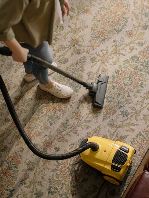 A person is vacuuming a patterned rug in a residential room using a yellow and black industrial-style carpet cleaner. The individual is dressed in casual clothing, including beige shoes and jeans, and