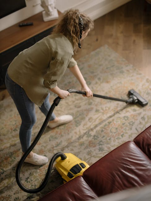 A person is vacuuming a patterned rug in a residential room using a yellow and black industrial-style carpet cleaner. The individual is dressed in casual clothing, including beige shoes and jeans, and