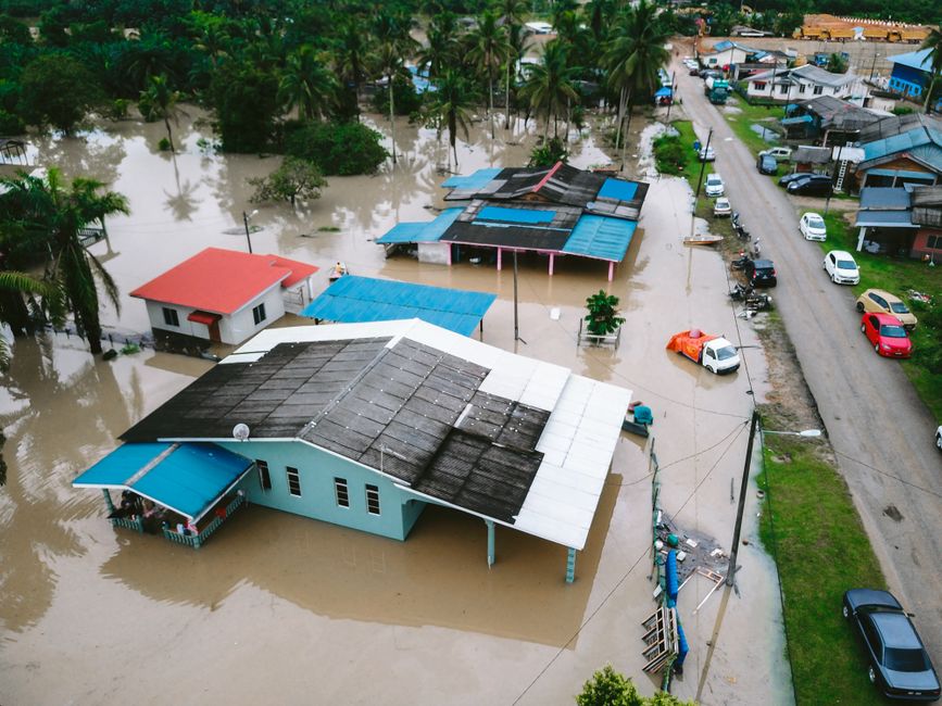 Aerial view of a residential area during a flood, showing multiple houses with their ground floors submerged in muddy water. The houses are constructed with various materials, including wood and concr