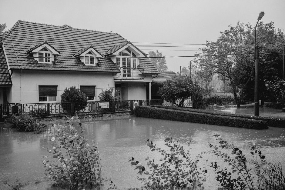 Aerial view of a residential area during a flood, showing multiple houses with their ground floors submerged in muddy water. The houses are constructed with various materials, including wood and concr